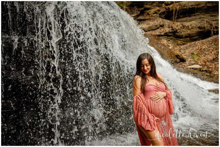 Pregnant woman holding belly standing next to waterfall.