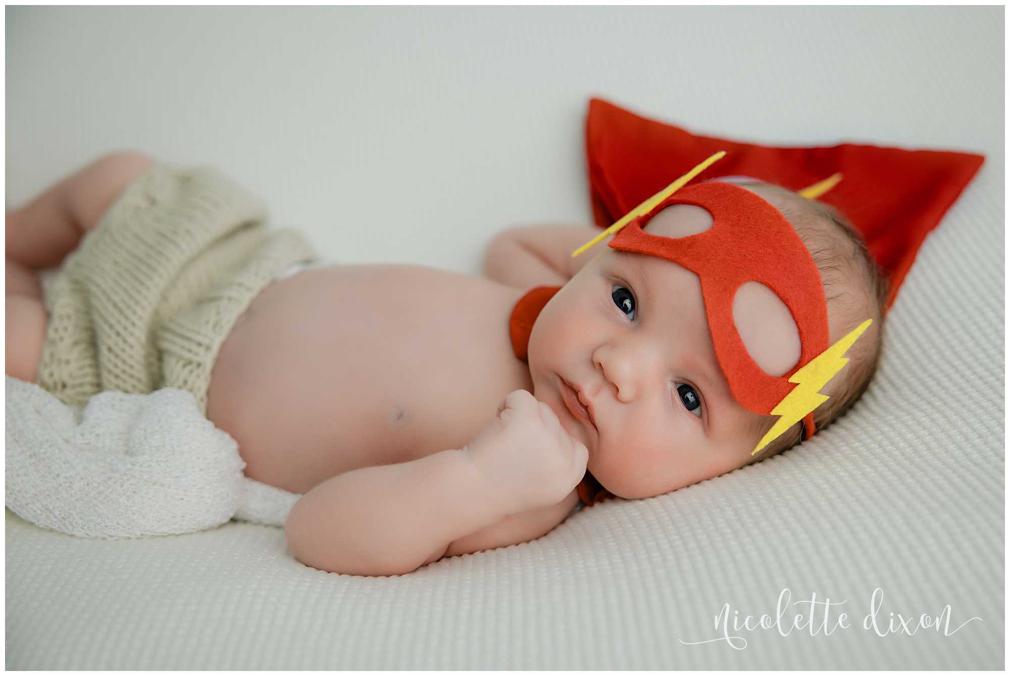 Newborn baby boy wearing flash costume while laying on white blanket in studio in North Fayette Township near Pittsburgh.